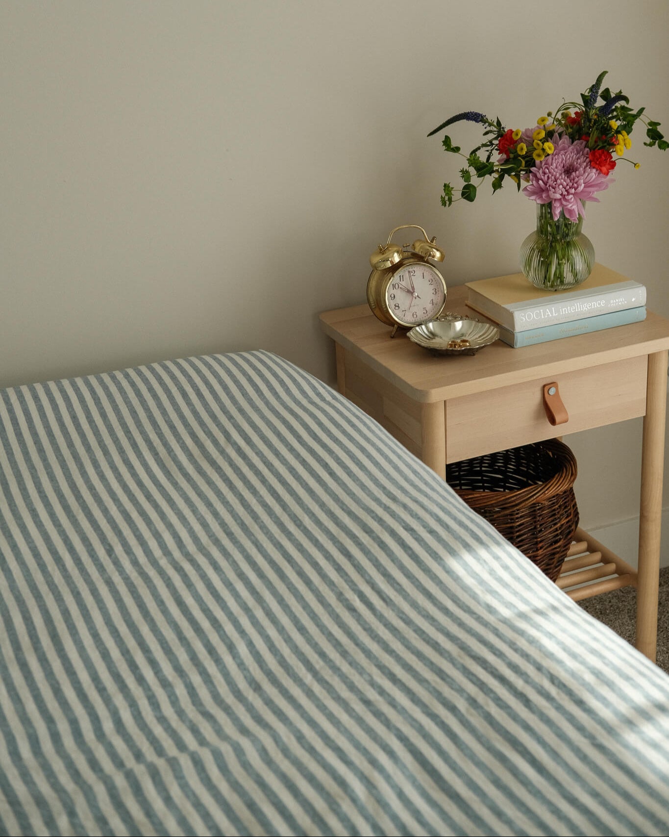 Bed with striped bedding next to a wooden nightstand with a clock, books, and flowers.