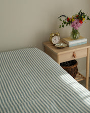 Bed with striped bedding next to a wooden nightstand with a clock, books, and flowers.