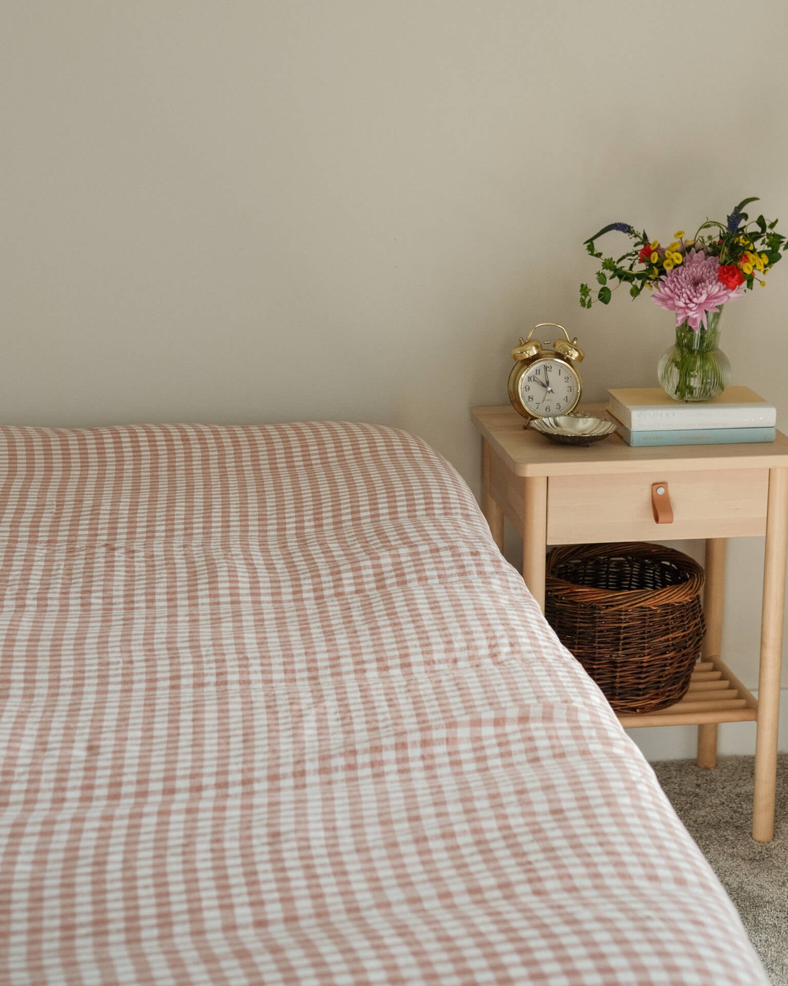 Bed with checkered bedding next to a wooden nightstand with a clock and flowers.